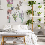 Real photo of white bedroom interior with many fresh plants, king-size bed, material painting with floral pattern and bench with books