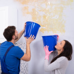 Young Woman And Repairman With Blue Bucket Collecting Water From Damaged Ceiling In Kitchen