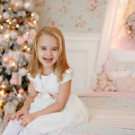 Very nice charming little girl blonde in white dress sitting on a child's bed and laughs loudly on the background of Christmas trees in bright interior of the house